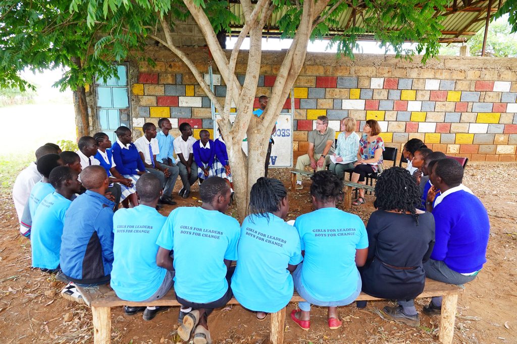 image members sit in a circle outdoors around a tree and hear from students from the girls for leaders program in Kenya